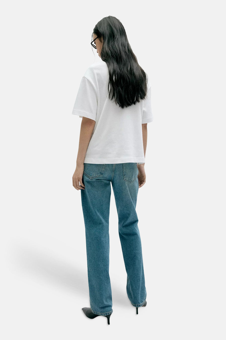 White short-sleeve t-shirt from back view, plain cotton, worn with blue jeans and black heels