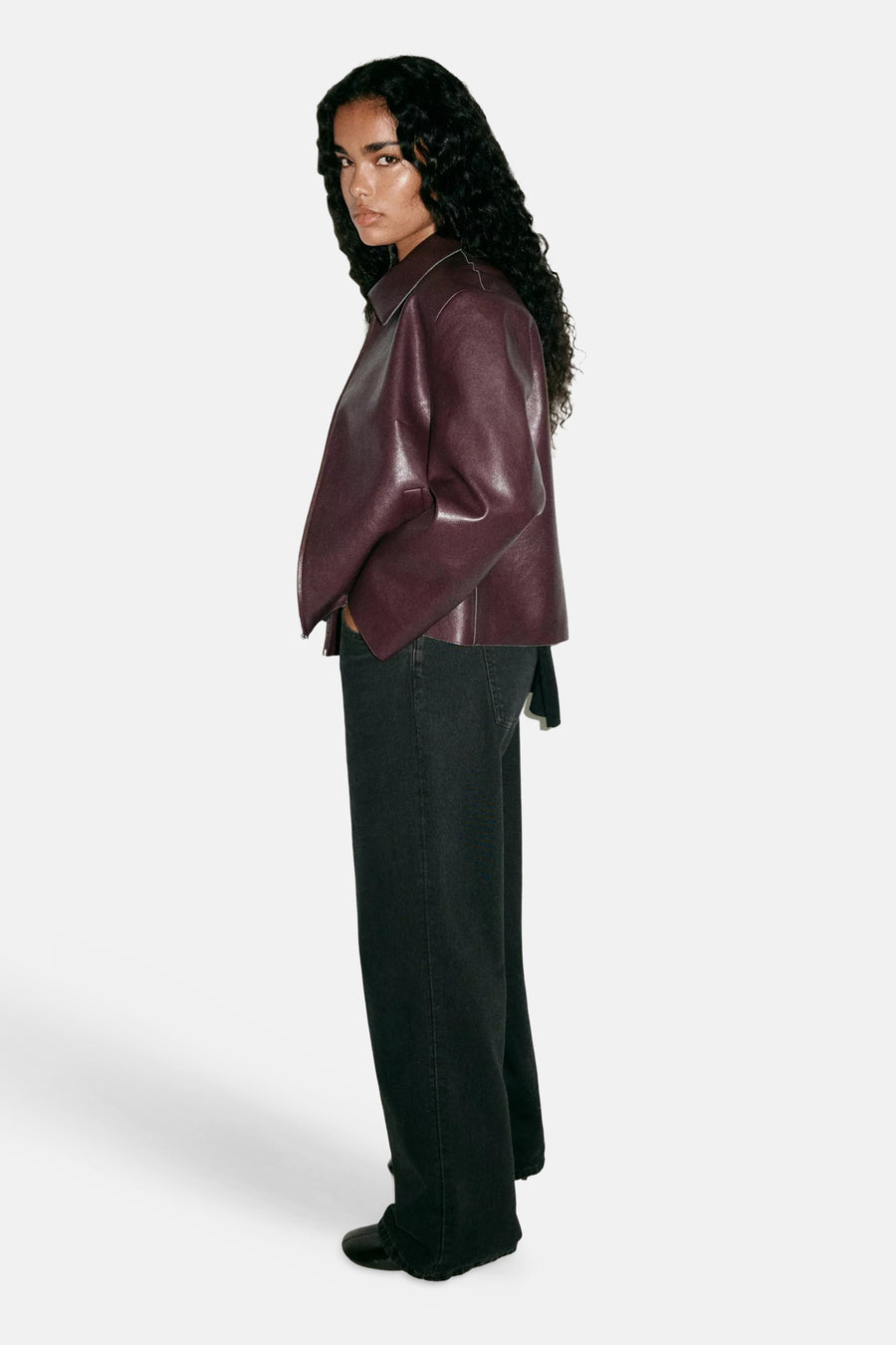 Bordeaux leather jacket with structured collar and side zipper, worn over black wide-leg jeans in side view