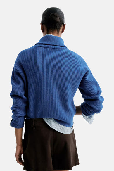 Blue ribbed turtleneck sweater back view, worn with rolled sleeves over a checkered shirt and dark shorts