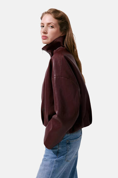 Bordeaux leather bomber jacket with high collar and relaxed fit, side view on model wearing blue jeans