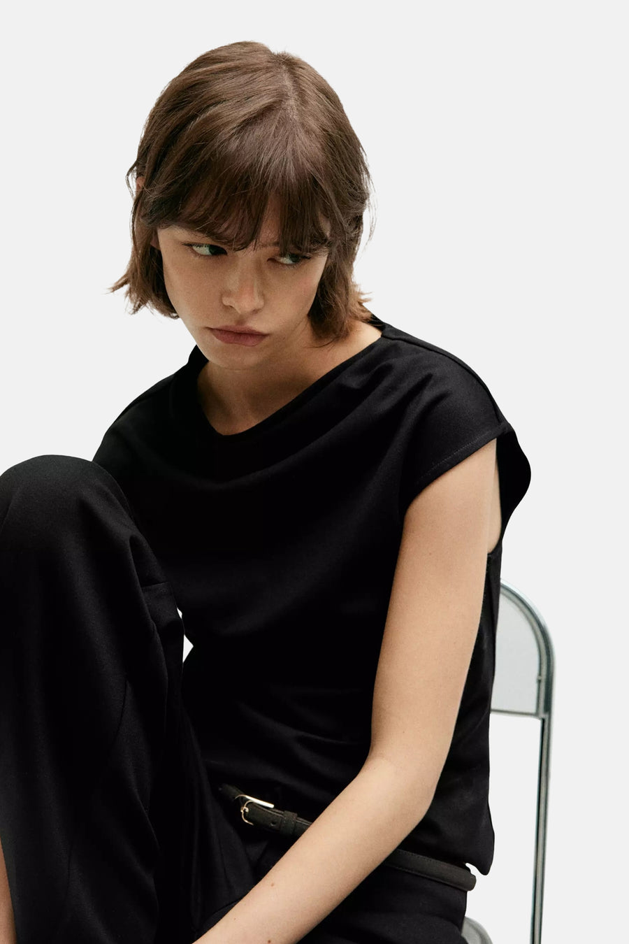 Black sleeveless top with draped neckline, worn by model in lifestyle pose, paired with black pants and belt