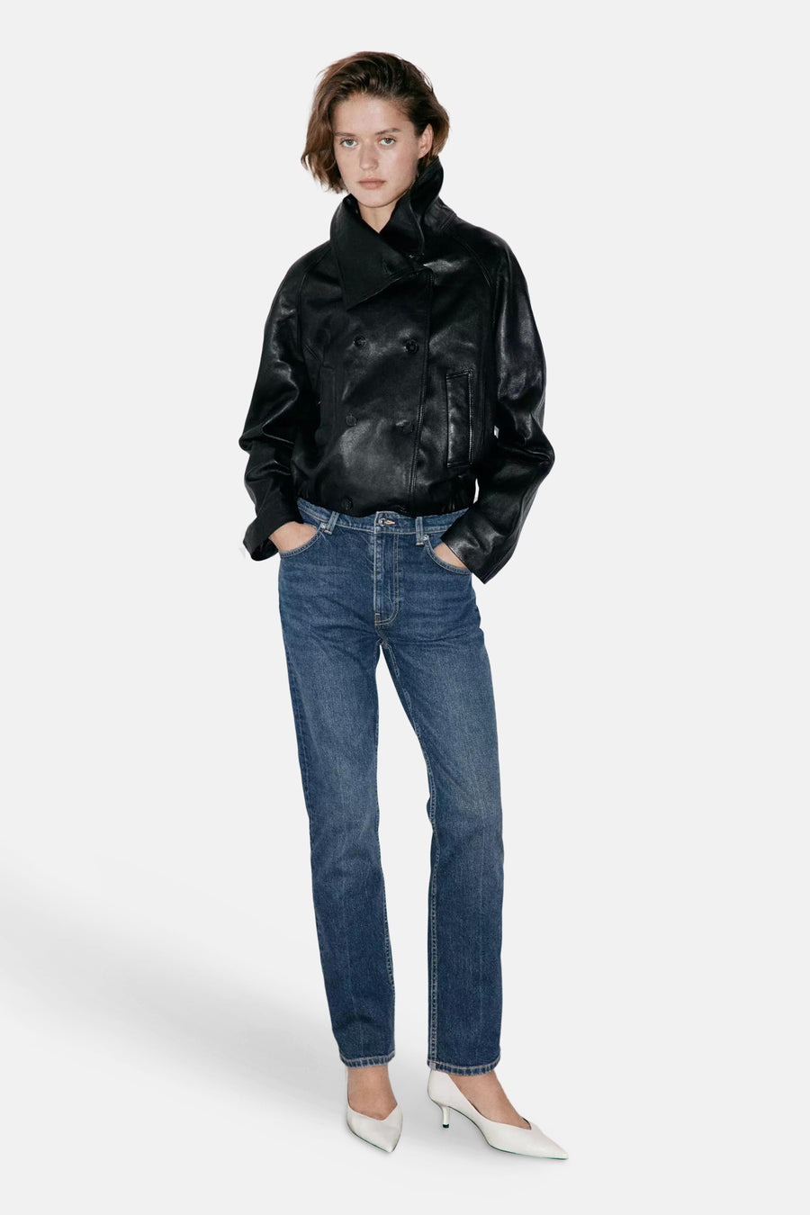 Jeans slim en denim bleu foncé par MANGO, coupe droite, portés avec une veste en cuir noir et escarpins blancs, vue de face.