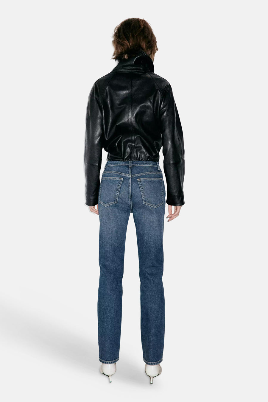 Jeans slim en denim bleu foncé par MANGO, vue arrière, avec poches carrées et coupe ajustée, portés avec une veste en cuir noir et escarpins blancs.
