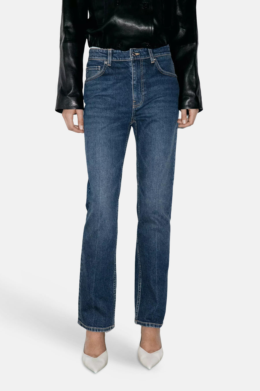 Jeans slim en denim bleu foncé par MANGO, coupe droite, portés avec une veste noire et des escarpins blancs, vue de face.