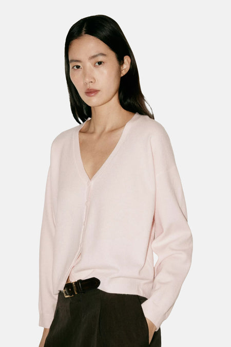 Cardigan rose en maille fine à col en V par MANGO, porté avec un pantalon noir et une ceinture en cuir, vue de face.