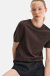 Brown short-sleeve T-shirt with floral beadwork, worn by model in lifestyle pose, paired with dark skirt.