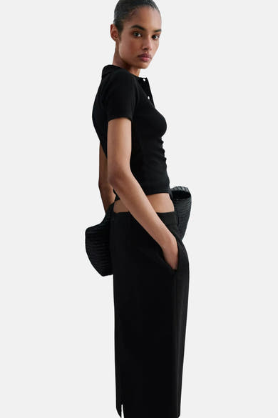 Black ribbed knit cardigan with button front, side view on model wearing black cropped top and wide-leg pants, holding woven bag.