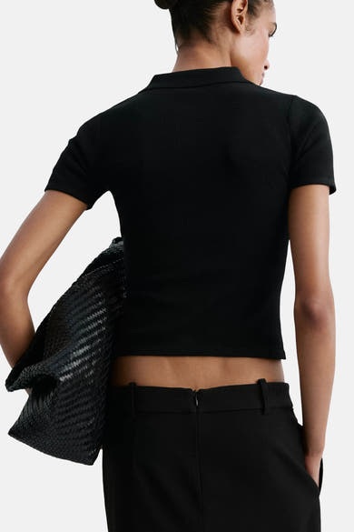 Black ribbed knit cardigan back view with short sleeves, worn over a cropped top and high-waisted pants, holding a woven black bag.