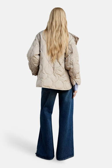 Beige quilted puffer jacket with oversized fit and hood, back view, worn with flared jeans and striped shirt cuff visible.
