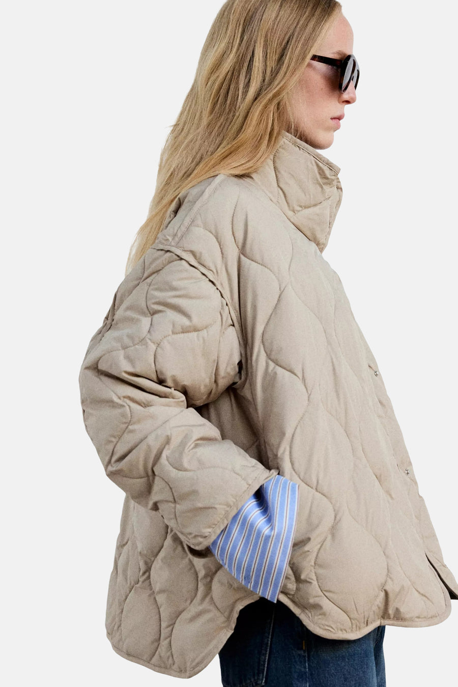 Beige quilted padded jacket with high collar and oversized fit, side view, worn with blue striped shirt sleeve visible