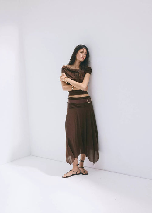 Brown wrap-style skirt with asymmetrical hem and belt loop detail, worn with matching top and sandals in full-length lifestyle pose.