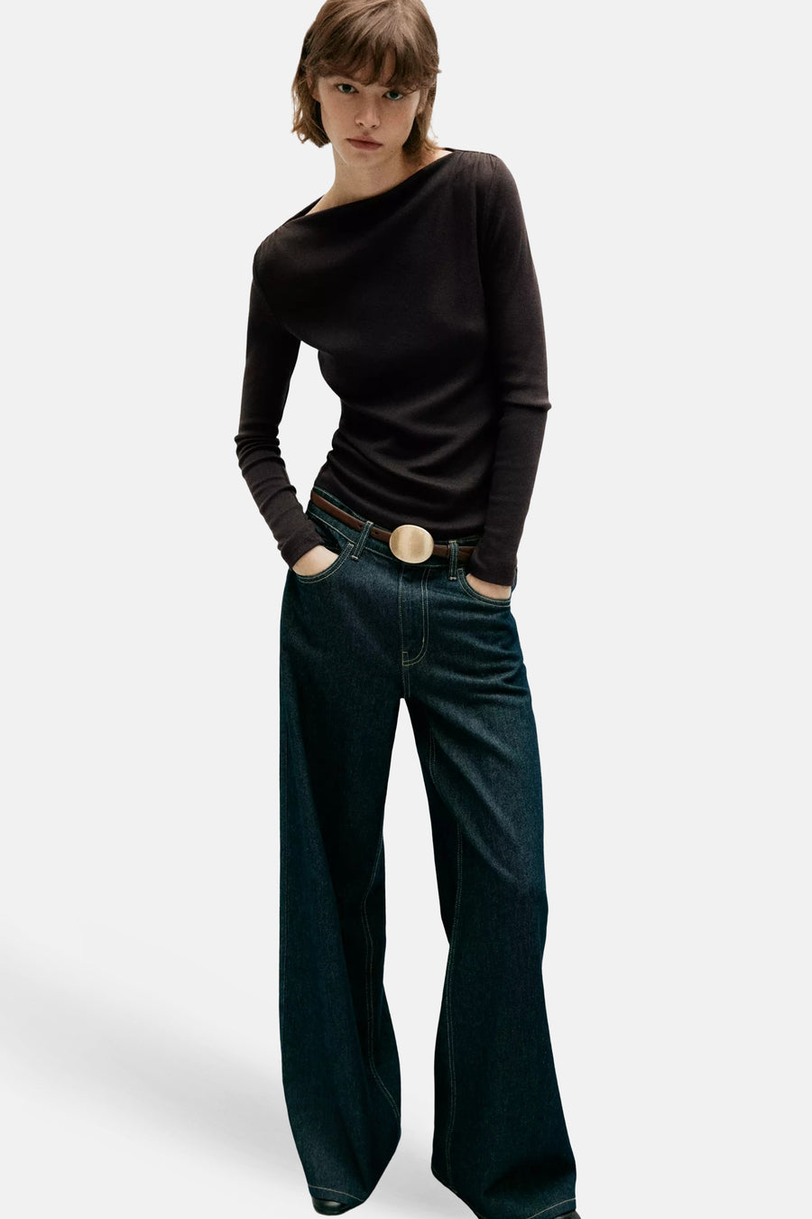 Brown long-sleeve top with asymmetrical neckline, worn with dark denim jeans and belt, full-length front view