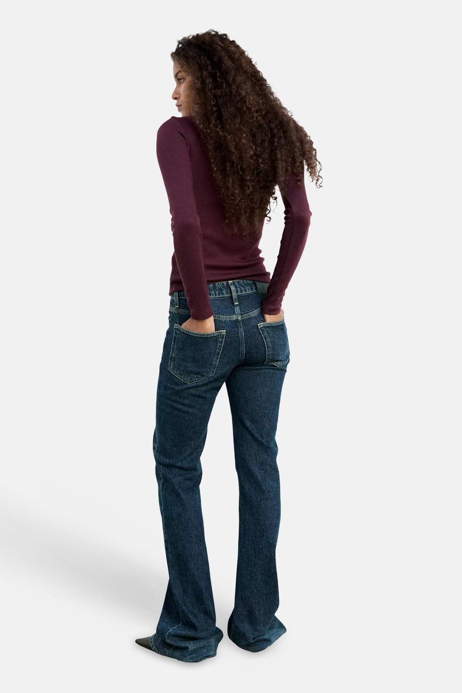 Bordeaux long-sleeve t-shirt by model in side view, paired with dark blue jeans, hands in pockets, curly hair flowing