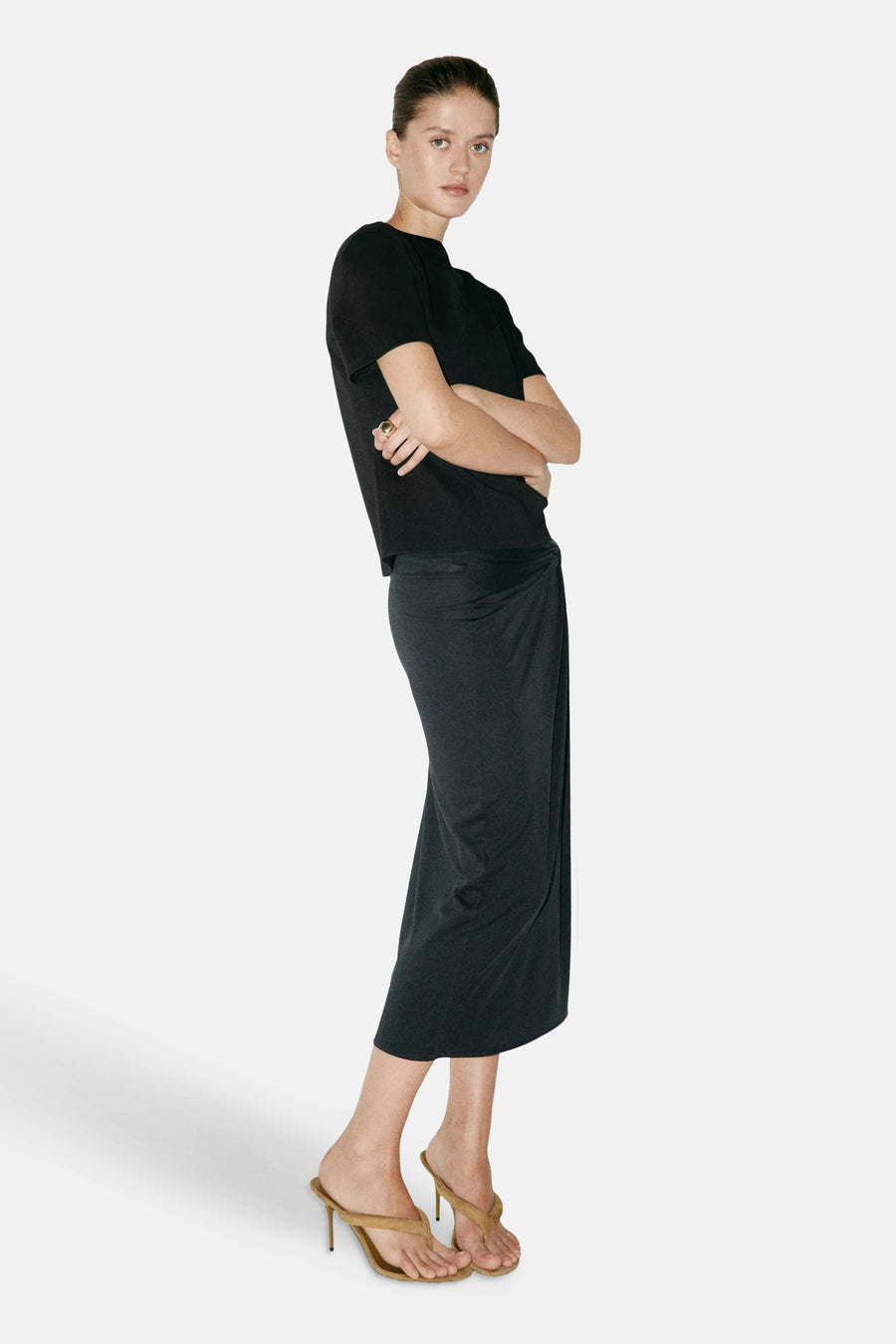 Black short-sleeve t-shirt with rounded neckline, worn by model in side view with black wrap skirt and tan heeled sandals.