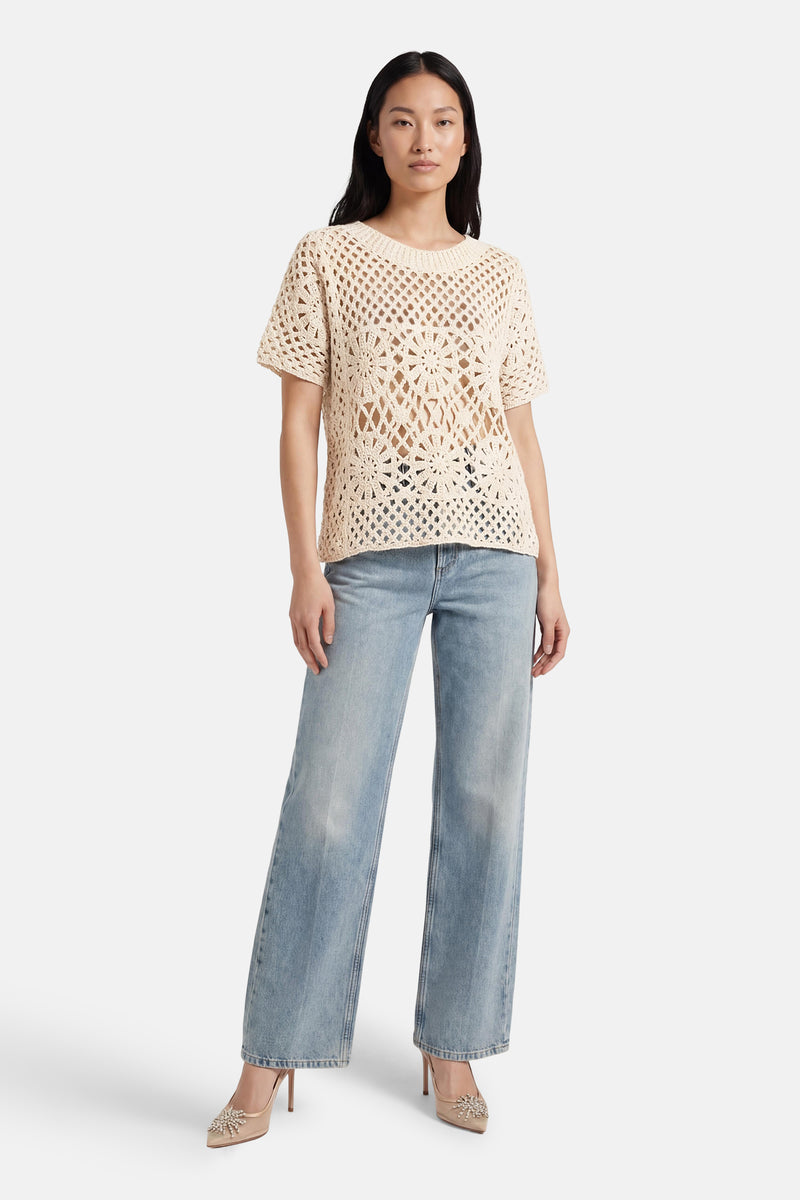 Pull beige à manches courtes de Terra di Siena, en crochet ajouré à motif floral, porté avec un jean ample et des escarpins ornés.