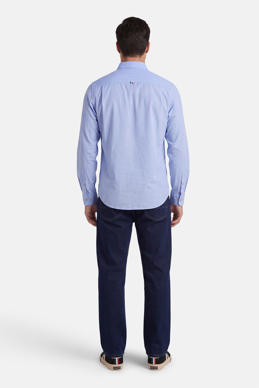 Chemise bleu ciel de DELAHAYE en tissu Oxford, vue de dos, avec un petit drapeau tricolore au col, portée avec un jean foncé et des baskets.