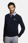 Polo bleu à manches longues de DELAHAYE, avec col bleu clair et logo rouge sur la poitrine, porté avec un pantalon rayé.
