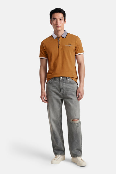 Polo brun de DELAHAYE, avec col à motif géométrique, poche poitrine et bordure blanche aux manches, porté avec un jean gris déchiré et des baskets.