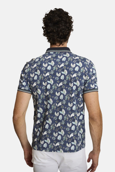 Polo bleu de DELAHAYE, imprimé avec motifs de feuilles, vu de dos, porté avec un pantalon blanc.
