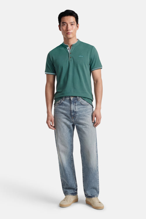 T-shirt bleu à manches courtes de DELAHAYE, avec patte de boutonnage et logo discret, porté avec un jean délavé et des baskets beiges.