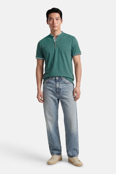 T-shirt bleu à manches courtes de DELAHAYE, avec patte de boutonnage et logo discret, porté avec un jean délavé et des baskets beiges.