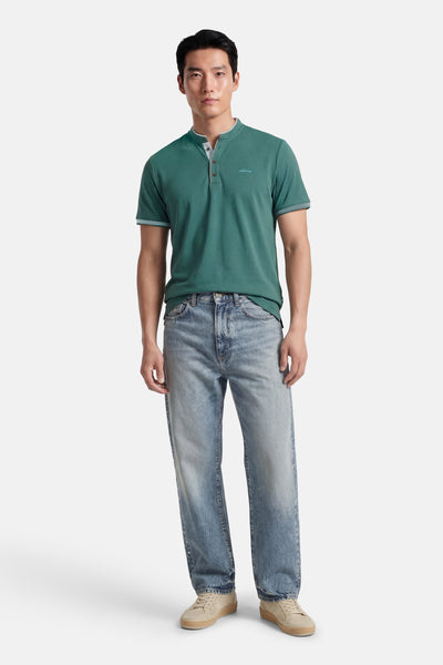 T-shirt bleu à manches courtes de DELAHAYE, avec patte de boutonnage et logo discret, porté avec un jean délavé et des baskets beiges.