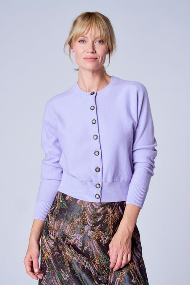 Gilet violet de TERRE BLEUE, avec des boutons dorés, porté avec une jupe à motifs.