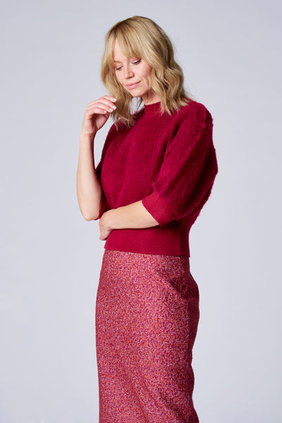 Pull bordeaux de TERRE BLEUE en maille duveteuse, porté avec une jupe rouge texturée et une frange blonde.
