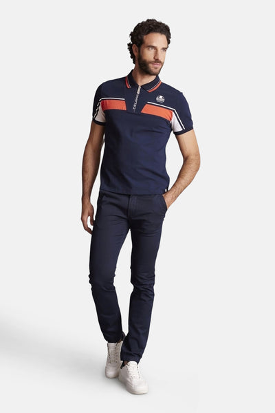 Polo bleu à manches courtes de DELAHAYE, avec rayures orange et blanches sur la poitrine et les manches, porté avec un pantalon bleu marine et des baskets blanches.
