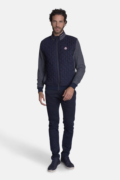 Gilet bleu marine matelassé de DELAHAYE, avec manches longues grises, porté sur un jean bleu foncé et des baskets marine.