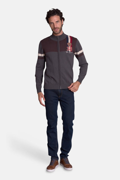 Brown knit gilet with zip front and striped sleeves, featuring embroidered "38 The Legend" patch on chest, worn by model in full-length lifestyle view.