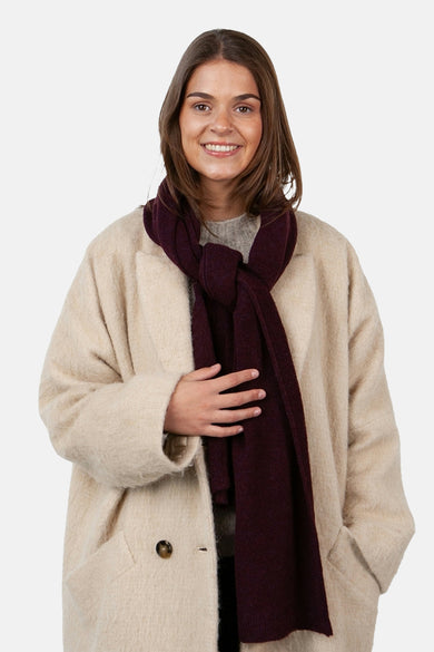 Écharpe rouge bordeaux de BARTS, nouée autour du cou, portée avec un manteau beige texturé.