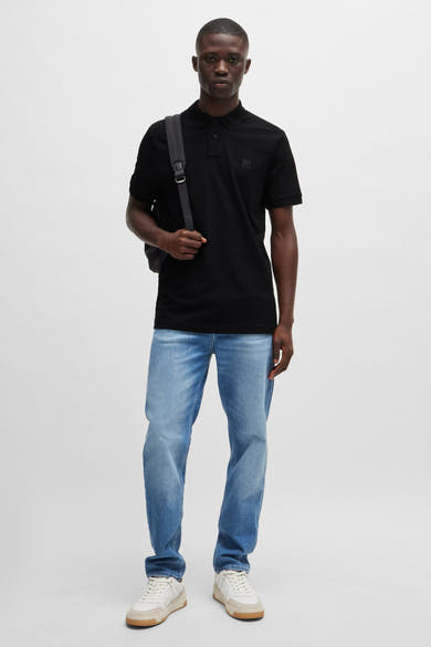 Zwart poloshirt met korte mouwen van BOSS ORANGE, gedragen met een blauwe jeans en crèmekleurige sneakers, met een rugzak.