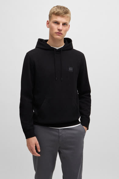 Sweat à capuche noir de BOSS ORANGE, avec cordon de serrage et poche kangourou, porté sur un pantalon gris.

