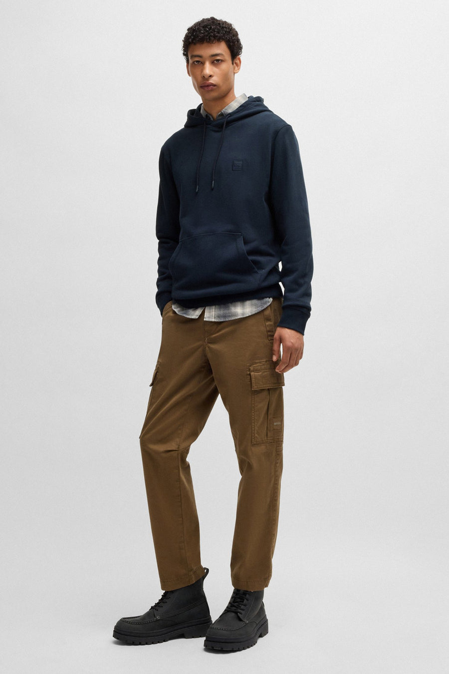 Blauwe BOSS ORANGE hoodie, gedragen over een geruit overhemd en een bruine cargobroek, met zwarte laarzen.