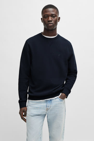 Sweat bleu de chez BOSS ORANGE, avec un logo discret et porté avec un t-shirt blanc et un jean délavé.