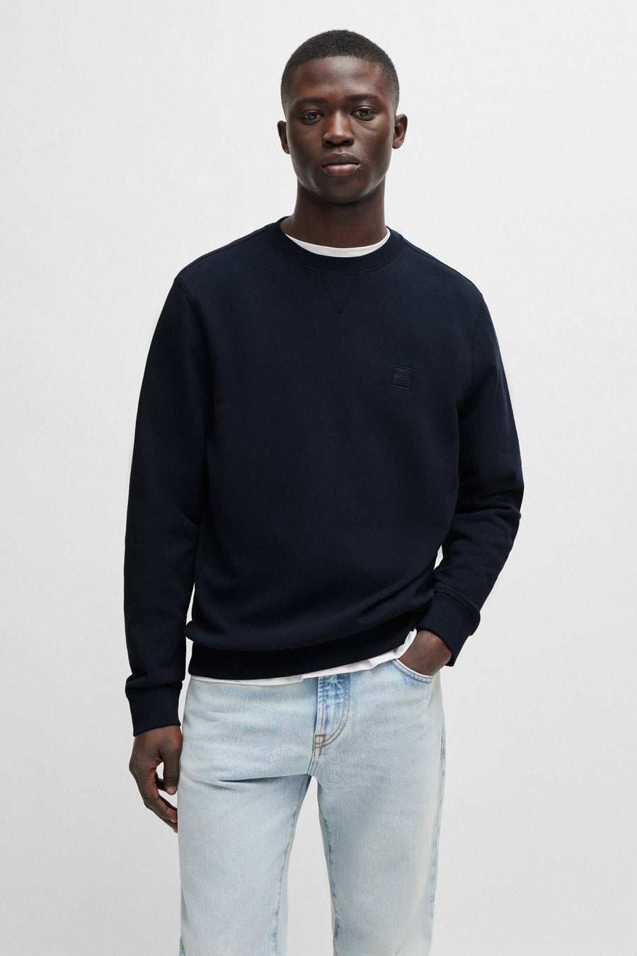 Sweat bleu de chez BOSS ORANGE, avec un logo discret et porté avec un t-shirt blanc et un jean délavé.