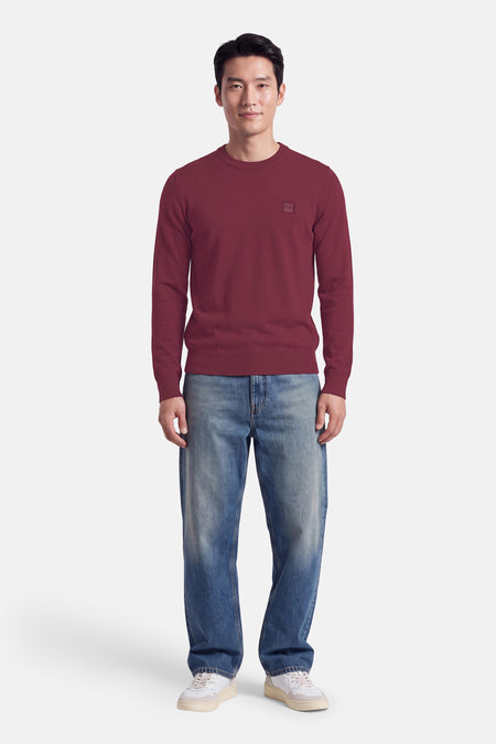 Pull col rond rouge de BOSS ORANGE, en maille fine, avec patch discret sur poitrine, porté avec jeans délavés et baskets blanches, vue frontale.