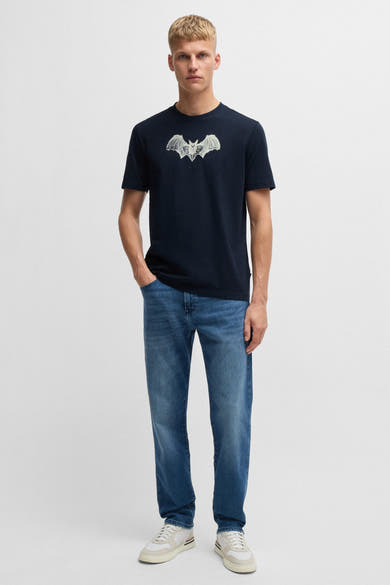 Blauw T-shirt met korte mouwen van BOSS ORANGE, met een vleermuisprint op de voorkant, gedragen met een spijkerbroek en sneakers.