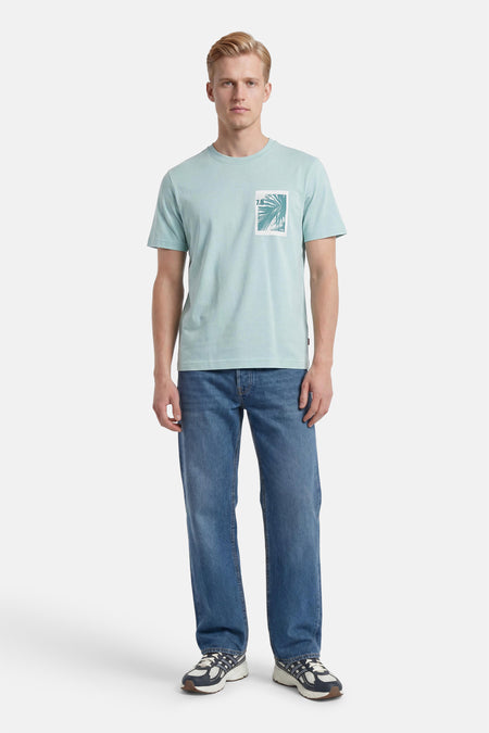 Turquoise T-shirt van BOSS ORANGE met een rechthoekige grafische palmbladprint, gedragen door een man met een blauwe spijkerbroek.