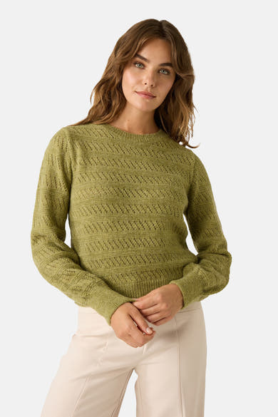 Groene trui met ronde hals van MORE & MORE, gedragen met een crèmekleurig jasje en een crèmekleurige broek, en een roze en beige geruite sjaal.