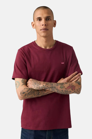 Bordeaux T-shirt met korte mouwen van Levi's, gedragen met een blauwe jeans en een subtiele ketting, armen over elkaar geslagen waardoor tatoeages zichtbaar zijn.