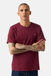 Bordeaux T-shirt met korte mouwen van Levi's, gedragen met een blauwe jeans en een subtiele ketting, armen over elkaar geslagen waardoor tatoeages zichtbaar zijn.