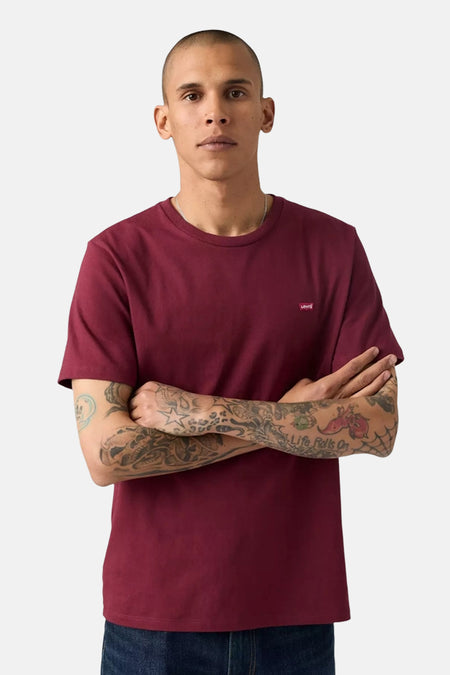 T-shirt bordeaux à manches courtes de Levi's, porté avec un jean bleu et un collier discret, les bras croisés révélant des tatouages.