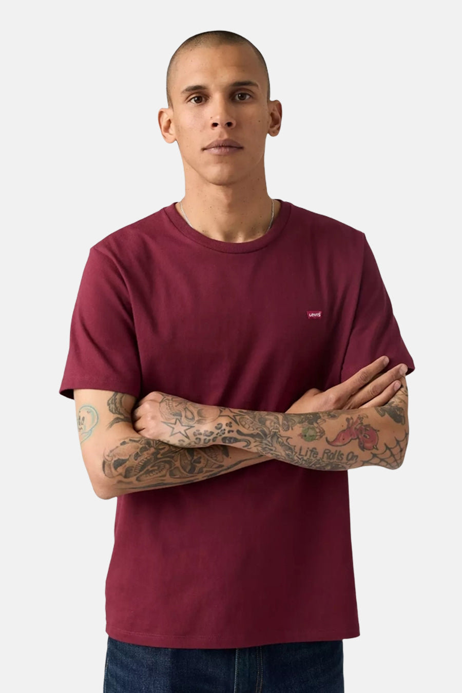 Bordeaux T-shirt met korte mouwen van Levi's, gedragen met een blauwe jeans en een subtiele ketting, armen over elkaar geslagen waardoor tatoeages zichtbaar zijn.