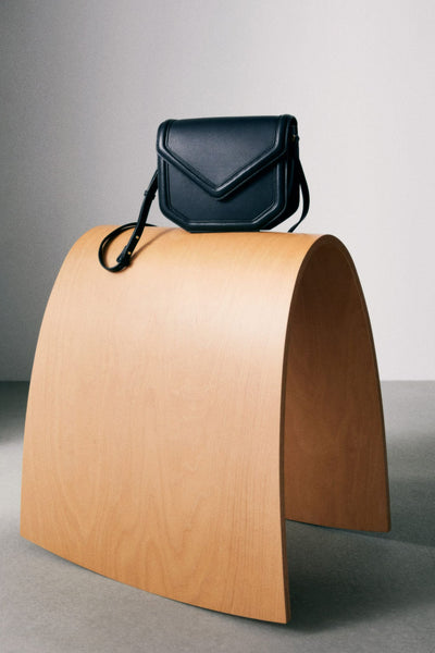 Black leather crossbody bag with flap closure and adjustable strap, displayed on wooden arch stand, front view