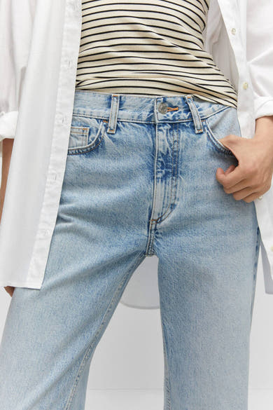 Jean droit bleu de MANGO, délavé avec une main dans la poche, porté avec un t-shirt rayé et une chemise blanche.
