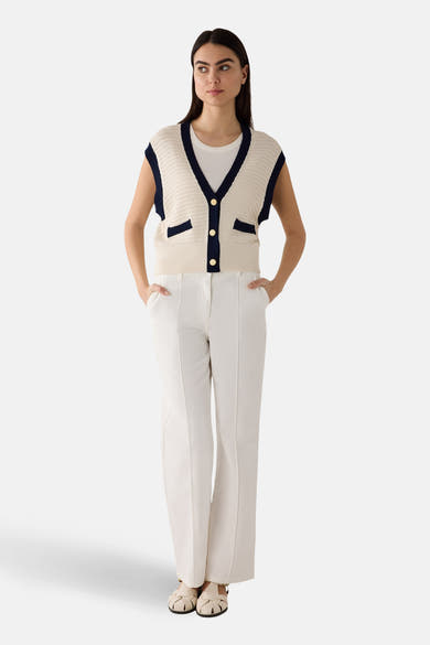 Gilet blanc en maille ajourée de More & More, avec bordures bleu marine et boutons dorés, porté sur un t-shirt blanc et un pantalon blanc.
