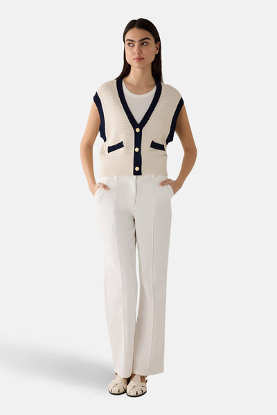 Gilet blanc en maille ajourée de More & More, avec bordures bleu marine et boutons dorés, porté sur un t-shirt blanc et un pantalon blanc.
