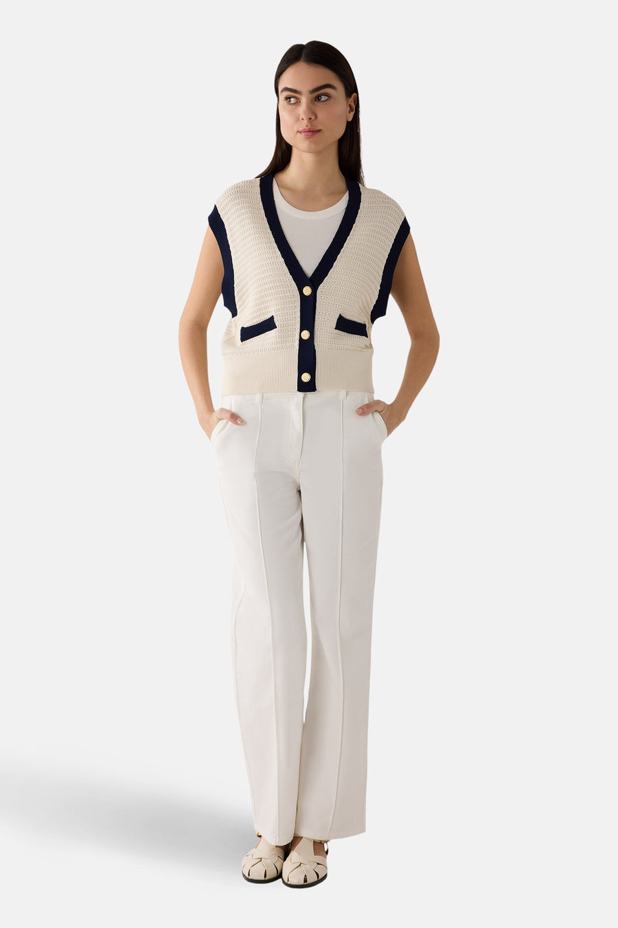 Gilet blanc en maille ajourée de More & More, avec bordures bleu marine et boutons dorés, porté sur un t-shirt blanc et un pantalon blanc.
